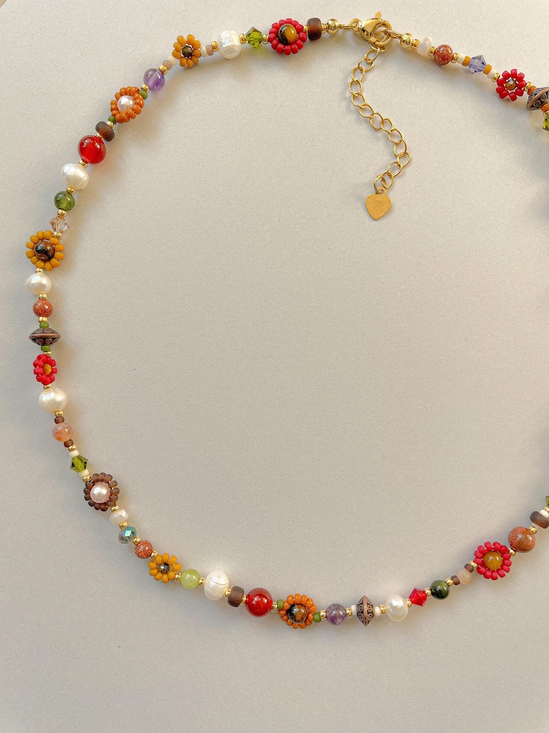 May include: A colorful beaded necklace featuring a variety of beads in shades of red, yellow, green, and brown. The necklace includes flower-shaped beads, pearls, and a gold-colored chain with a heart-shaped charm.