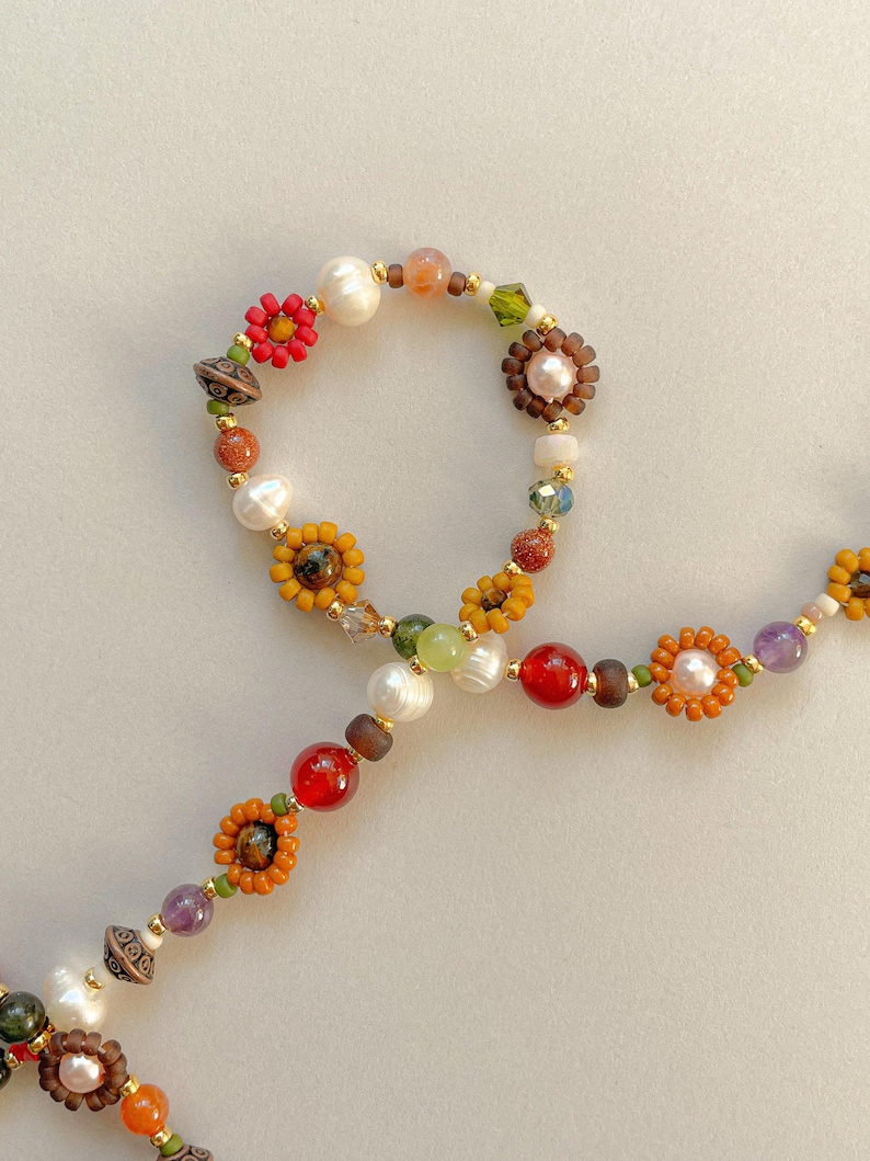 May include: A colorful beaded necklace featuring a variety of beads, including pearls, red and orange beads, and flower-shaped accents. The necklace has a mix of gold, brown, and green beads, creating a vibrant and eye-catching design.