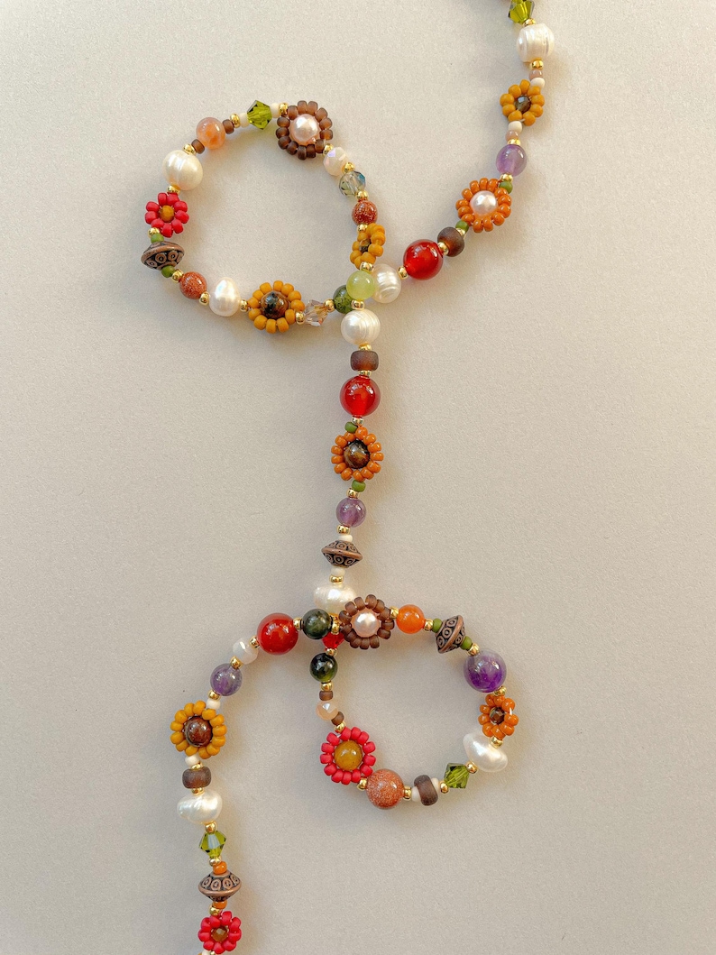 May include: A handcrafted necklace featuring a unique design with colorful beads, including pearls, red, orange, and purple gemstones, and flower-shaped accents. The necklace is made with gold-colored metal and has a delicate, artistic appearance.