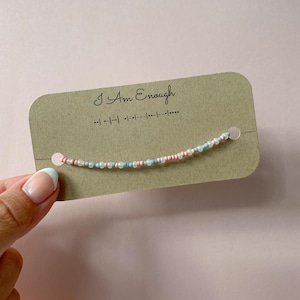 以下が含まれることがあります： ピンク、ブルー、クリアのビーズが付いた繊細なビーズブレスレットが、茶色のカードに表示されています。カードには筆記体で「I Am Enough」と書かれており、その下にモールス信号があります。ブレスレットはフレンチマニキュアをした手で持たれています。