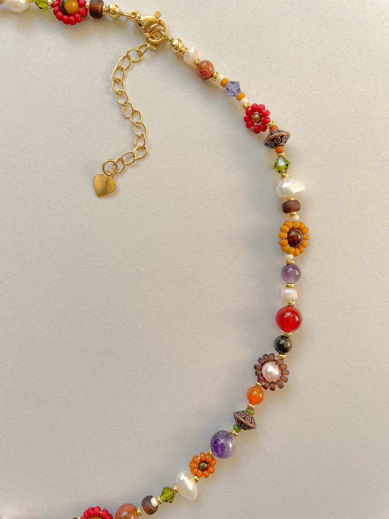 May include: A colorful beaded necklace with a gold chain and heart-shaped clasp. The necklace features a variety of beads, including red flower-shaped beads, pearls, and gemstones in shades of orange, purple, and green. The necklace is on a light background.