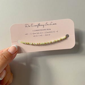 May include: A beaded bracelet with a light green and gold color scheme is displayed on a pink card. The card has the text "Do Everything In Love" and "I Corinthians 16:14". The bracelet is made of small beads and is strung together.