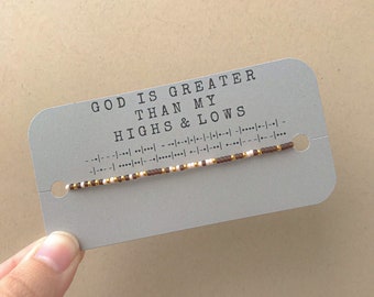 May include: A grey card with the text "GOD IS GREATER THAN MY HIGHS & LOWS" and a brown, white and gold beaded bracelet.