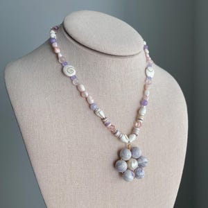 Op de afbeelding: Een kralenketting met een bloemvormige hanger bestaande uit zes ronde, lichtpaarse gemarmerde kralen rondom een centrale witte kraal. Het snoer bestaat uit kleine witte schelpjes, doorschijnende lichtpaarse en zachtroze kralen.