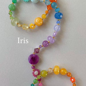 May include: A multi-colored beaded necklace featuring a variety of round, faceted, and flower-shaped beads in shades of red, orange, yellow, green, blue, purple, and pink, accented with small gold-tone spacers and the word Iris printed in white.