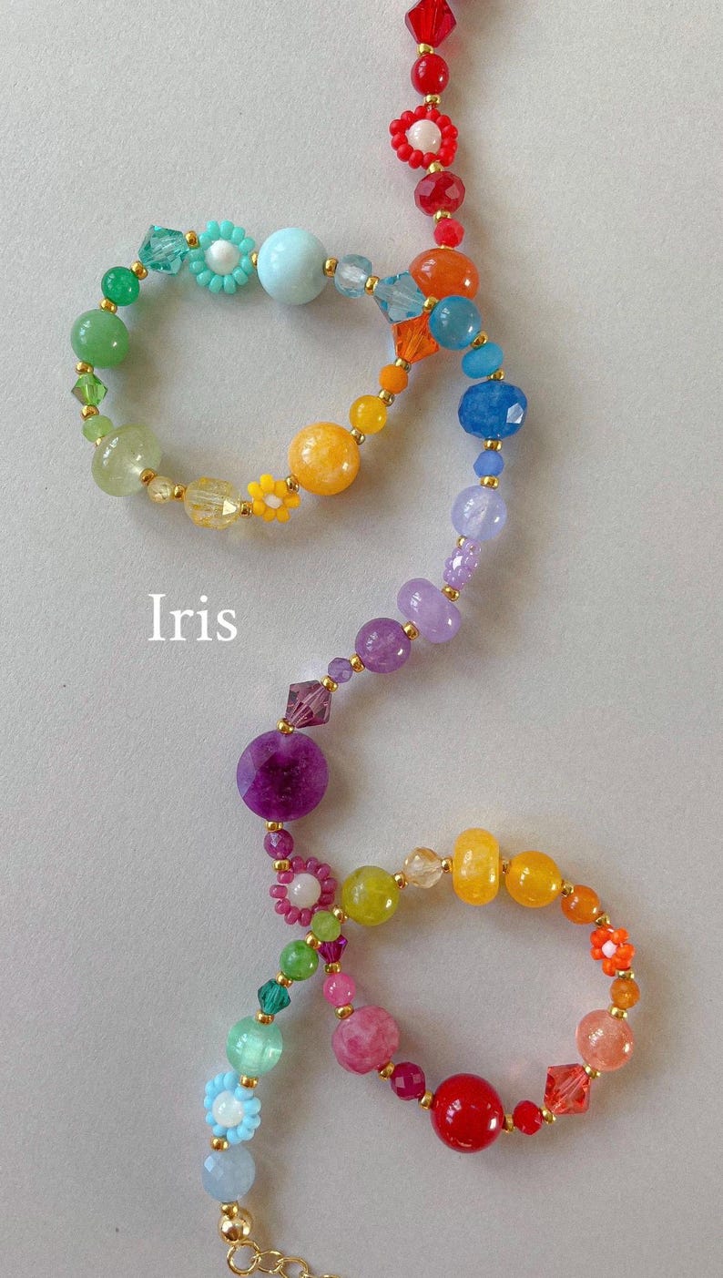 May include: A multi-colored beaded necklace featuring a variety of round, faceted, and flower-shaped beads in shades of red, orange, yellow, green, blue, purple, and pink, accented with small gold-tone spacers and the word Iris printed in white.