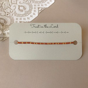 Puede incluir: Un brazalete de cuentas con cuentas de color óxido y doradas se muestra en una tarjeta que dice "Trust in the Lord". La tarjeta también presenta código Morse. El brazalete está ensartado en un cordón fino y está centrado en la tarjeta.