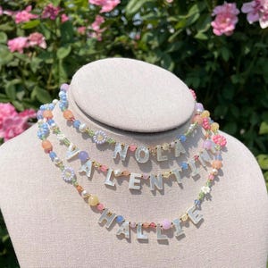 May include: Three layered necklaces featuring colorful beads in shades of pink, blue, yellow, and green, interspersed with small gold-tone spacers. Each strand displays white letter beads spelling out NOLA, VALENTINA, and HALLIE respectively.