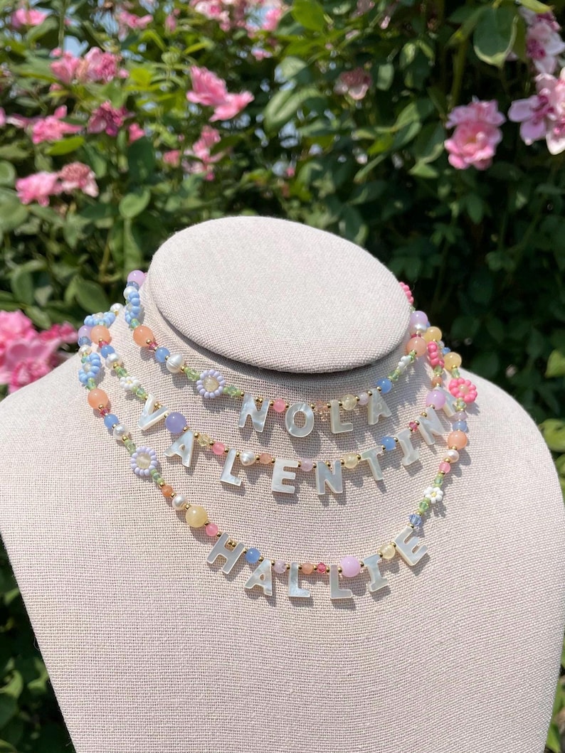 May include: Three layered necklaces featuring colorful beads in shades of pink, blue, yellow, and green, interspersed with small gold-tone spacers. Each strand displays white letter beads spelling out NOLA, VALENTINA, and HALLIE respectively.