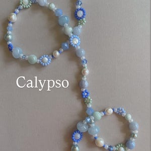 May include: A beaded necklace featuring a variety of light blue, pale green, and white beads, including small flower-shaped beads and round pearls. The word Calypso is printed in white text on the left side of the necklace arrangement.