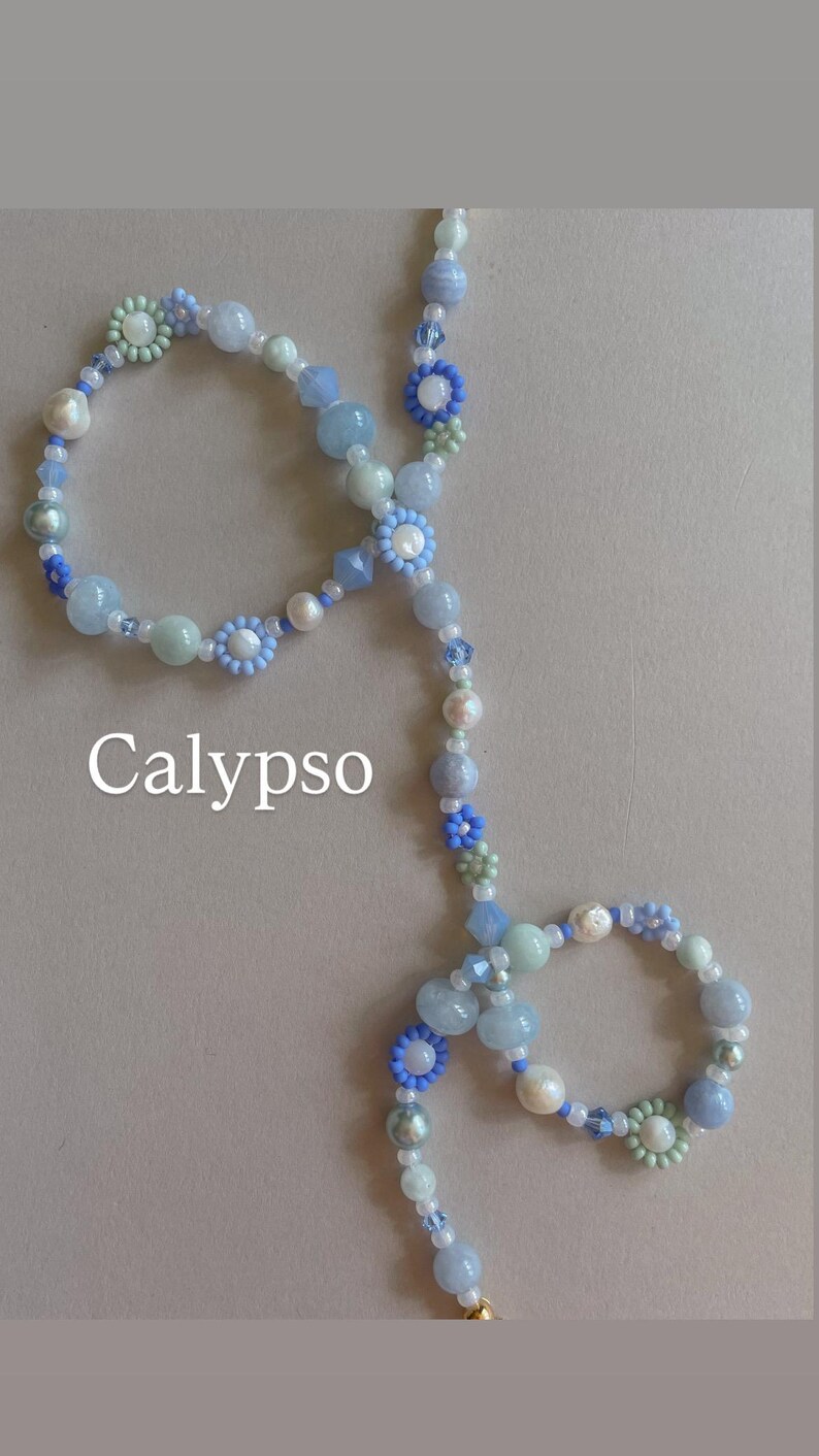 May include: A beaded necklace featuring a variety of light blue, pale green, and white beads, including small flower-shaped beads and round pearls. The word Calypso is printed in white text on the left side of the necklace arrangement.
