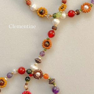 May include: A beaded necklace featuring the word Clementine, composed of multicolored glass beads, white freshwater pearls, and small flower-shaped bead clusters in shades of red, orange, yellow, and purple, accented with metallic gold spacers.