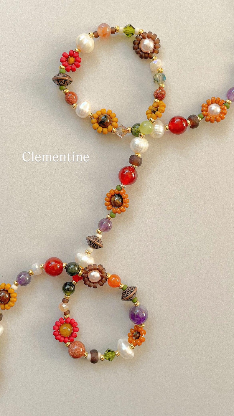 May include: A beaded necklace featuring the word Clementine, composed of multicolored glass beads, white freshwater pearls, and small flower-shaped bead clusters in shades of red, orange, yellow, and purple, accented with metallic gold spacers.