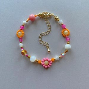 May include: A colorful beaded bracelet featuring a gold clasp and extender chain. The bracelet is adorned with orange, pink, white, and gold beads, including flower-shaped accents. The bracelet is approximately 7 inches long.