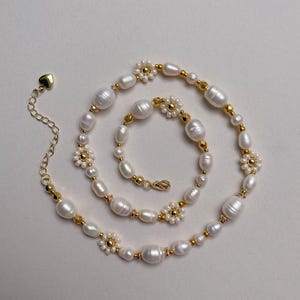 Peut inclure: Un collier délicat composé de perles d'eau douce blanches, de perles dorées et d'éléments en forme de marguerite. La chaîne dorée comprend un charm en forme de cœur et une fermeture à fermoir. Le bijou est présenté sur un fond blanc.