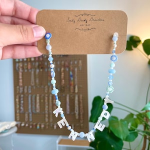 May include: A beaded necklace featuring light blue, white, and clear beads with white letter beads spelling KENNEDY. The necklace is attached to a brown cardboard display card printed with the text Seedy Beady Bracelets AND THINGS.