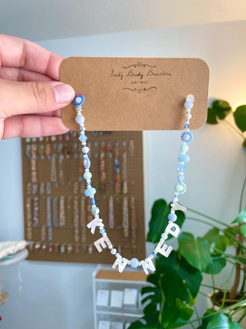 May include: A beaded necklace featuring light blue, white, and clear beads with white letter beads spelling KENNEDY. The necklace is attached to a brown cardboard display card printed with the text Seedy Beady Bracelets AND THINGS.