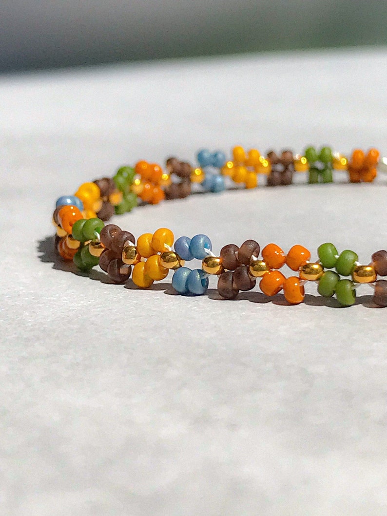 May include: A close-up of a beaded bracelet featuring a multi-coloured pattern. The bracelet is made with small, round beads in shades of brown, orange, yellow, blue, and green. The beads are strung on a thin wire and are arranged in a repeating pattern.