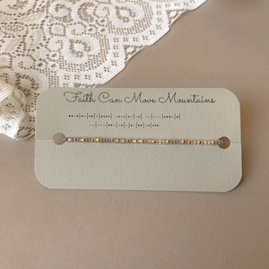 May include: A beaded bracelet with gold and gray beads is displayed on a light gray card. The card reads "Faith Can Move Mountains" in script, with a line of Morse code below. The bracelet is on a neutral background.