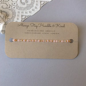 May include: A pink and gold beaded bracelet with the text "Always Stay Humble & Kind" on a brown card.