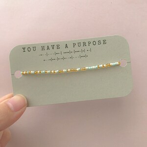 May include: A light green card with the text "YOU HAVE A PURPOSE" printed in black. A thin beaded bracelet with white, light blue, and gold beads is wrapped around the card.