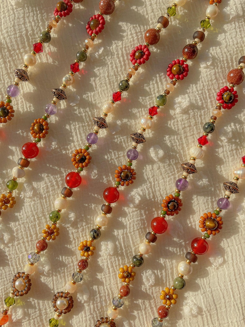 May include: Close-up of several strands of beaded jewelry. The strands feature a variety of beads in colors like red, orange, brown, green, and purple. Some beads are floral-shaped, and others are round or faceted. The background is a textured white fabric.