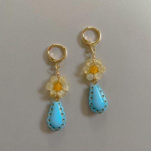 以下が含まれることがあります： 黄色と白のビーズでできた花の形をしたビーズと、青い涙の形をしたビーズが付いた、金色のぶら下がり式のイヤリングのペアです。
