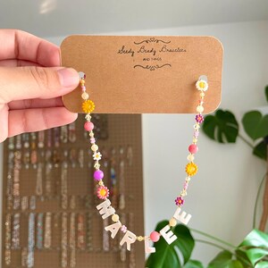 May include: A beaded necklace on a brown cardboard display card labeled Seedy Beady Bracelets AND THINGS. The necklace features colorful round beads, small flower-shaped beads in yellow, pink, and white, and white letter beads spelling MARLEE.