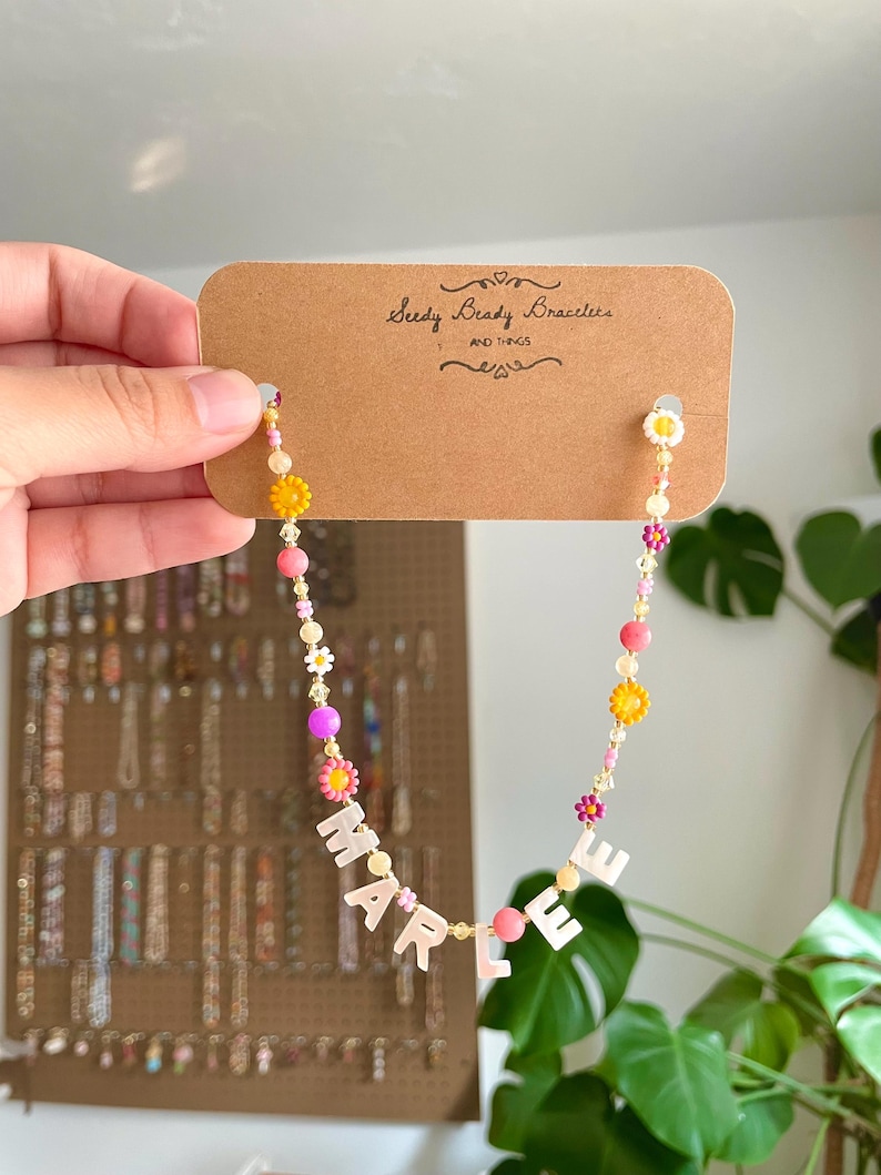 May include: A beaded necklace on a brown cardboard display card labeled Seedy Beady Bracelets AND THINGS. The necklace features colorful round beads, small flower-shaped beads in yellow, pink, and white, and white letter beads spelling MARLEE.