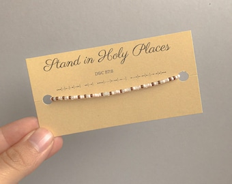 Op de afbeelding: Een delicate kralenarmband met witte, bruine en lichtroze kralen wordt getoond op een bruine kaart. De kaart heeft de tekst "Stand in Holy Places". De armband wordt door een hand vastgehouden tegen een neutrale grijze achtergrond.