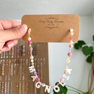 May include: A beaded necklace featuring white mother-of-pearl letter beads spelling MAGNOLIA, interspersed with pink, peach, purple, and yellow beads, including small flower-shaped beads, attached to a brown cardboard display card labeled Seedy Beady Bracelets.