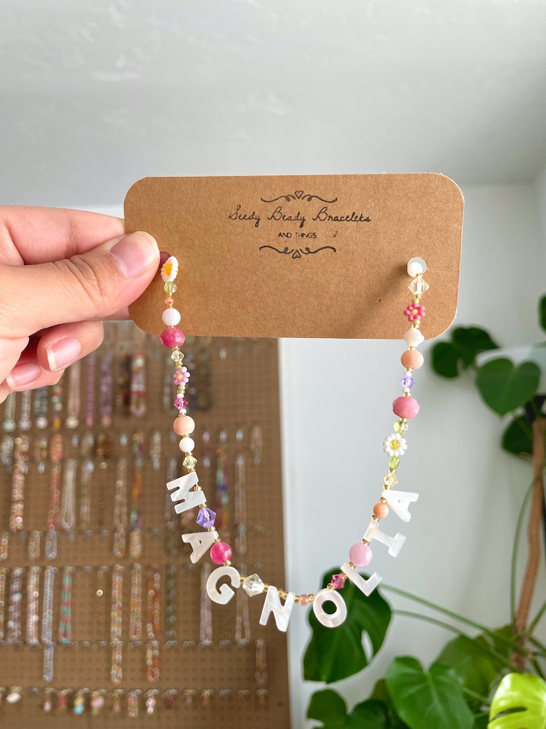 May include: A beaded necklace featuring white mother-of-pearl letter beads spelling MAGNOLIA, interspersed with pink, peach, purple, and yellow beads, including small flower-shaped beads, attached to a brown cardboard display card labeled Seedy Beady Bracelets.