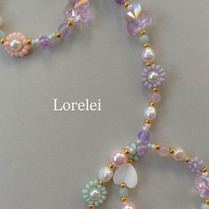 May include: A beaded necklace featuring pastel-colored pearls, translucent butterfly-shaped beads, flower-shaped beads with pearl centers, and white heart-shaped charms, all accented with small gold-tone spacers. The text Lorelei is visible.