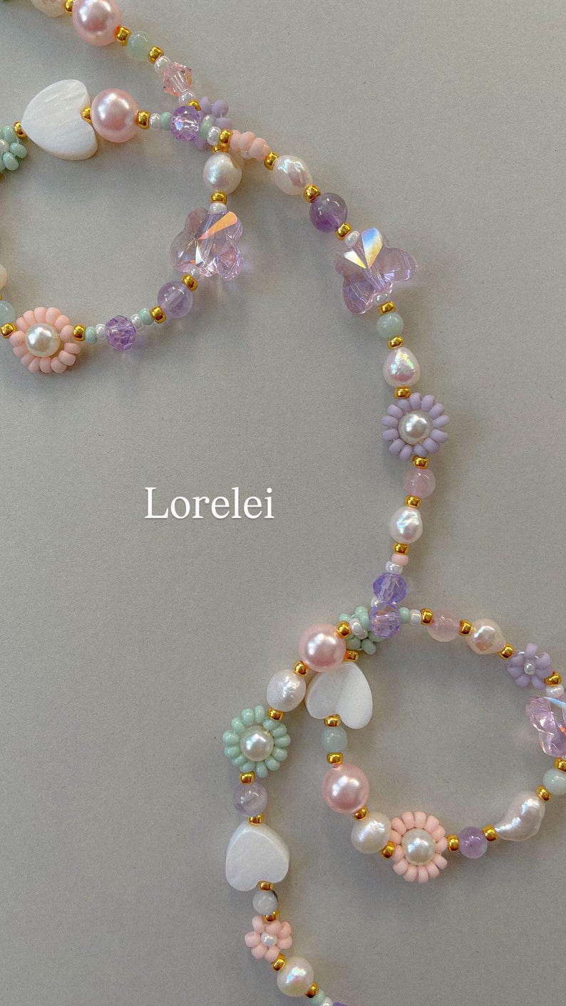 May include: A beaded necklace featuring pastel-colored pearls, translucent butterfly-shaped beads, flower-shaped beads with pearl centers, and white heart-shaped charms, all accented with small gold-tone spacers. The text Lorelei is visible.