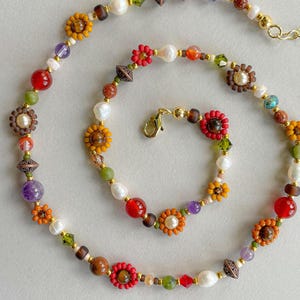 May include: A colorful beaded necklace featuring a variety of beads in shades of red, orange, yellow, green, purple, and white. The necklace includes flower-shaped beads, pearls, and other decorative elements, with a gold-colored clasp.