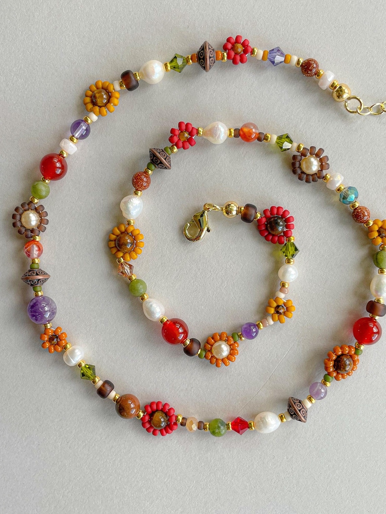 May include: A colorful beaded necklace featuring a variety of beads in shades of red, orange, yellow, green, purple, and white. The necklace includes flower-shaped beads, pearls, and other decorative elements, with a gold-colored clasp.