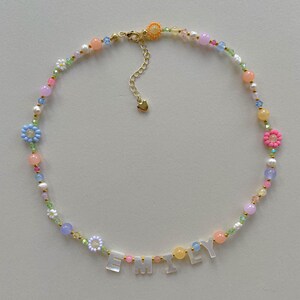 May include: A beaded necklace featuring a variety of colorful round beads, small flower-shaped beads, and white letter beads spelling the name EMILY. The necklace includes a gold-toned metal clasp and an adjustable chain with a small heart charm.
