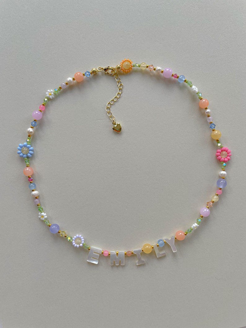 May include: A beaded necklace featuring a variety of colorful round beads, small flower-shaped beads, and white letter beads spelling the name EMILY. The necklace includes a gold-toned metal clasp and an adjustable chain with a small heart charm.