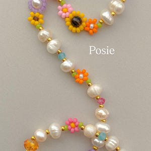 May include: A beaded necklace featuring white freshwater pearls, small colorful flower-shaped beads in yellow, orange, pink, and blue, and translucent faceted crystals in purple and amber, accented with gold-tone spacers and the text Posie.