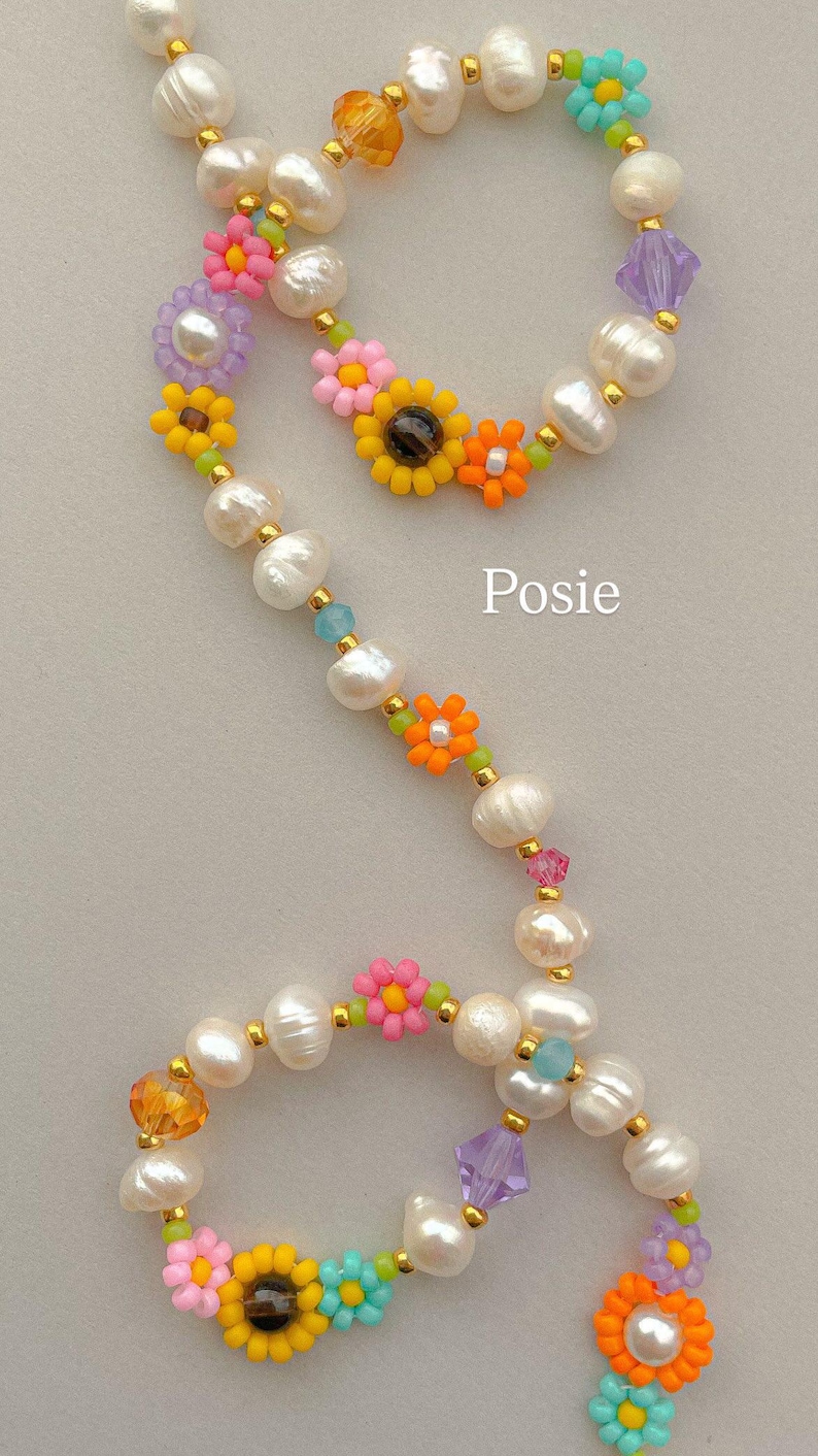 May include: A beaded necklace featuring white freshwater pearls, small colorful flower-shaped beads in yellow, orange, pink, and blue, and translucent faceted crystals in purple and amber, accented with gold-tone spacers and the text Posie.
