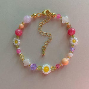 May include: A beaded bracelet featuring a mix of colorful beads, including pink, white, yellow, and purple. The bracelet has a gold-colored clasp and chain. The beads are arranged in a pattern with daisy and clover-shaped accents.
