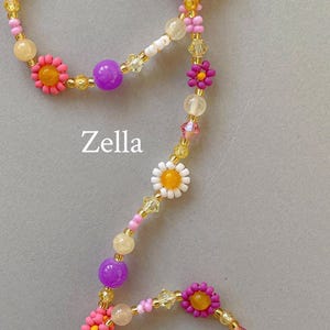 May include: A beaded necklace featuring the text Zella, composed of multi-colored floral-shaped beads in pink, orange, white, and purple, interspersed with translucent yellow, pink, and clear faceted crystal beads on a thin gold-toned wire.