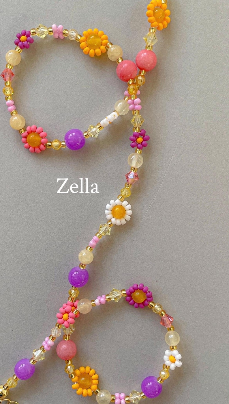 May include: A beaded necklace featuring the text Zella, composed of multi-colored floral-shaped beads in pink, orange, white, and purple, interspersed with translucent yellow, pink, and clear faceted crystal beads on a thin gold-toned wire.
