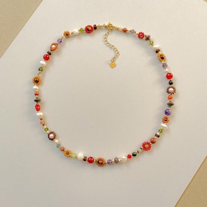 May include: A colorful beaded necklace featuring a variety of beads, including pearls, red, orange, and yellow flower-shaped beads, and other colorful gemstones. The necklace has a gold-colored clasp and chain.