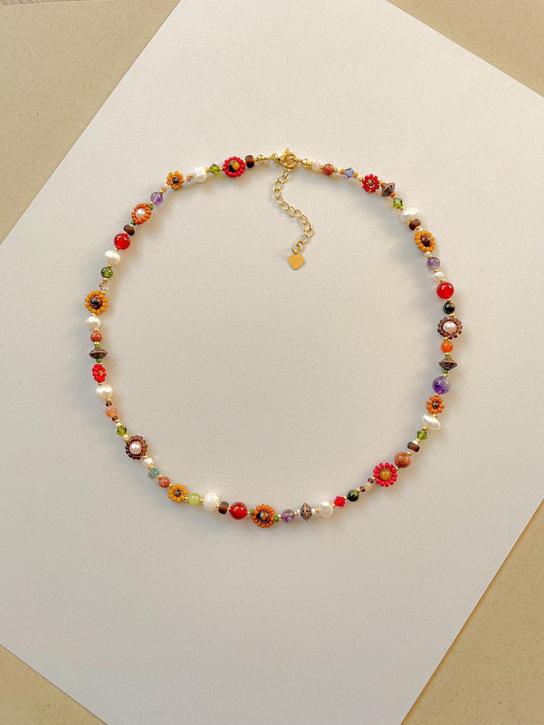 May include: A colorful beaded necklace featuring a variety of beads, including pearls, red, orange, and yellow flower-shaped beads, and other colorful gemstones. The necklace has a gold-colored clasp and chain.