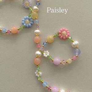 May include: A beaded necklace labeled Paisley featuring a variety of pastel-colored beads, including white pearls, translucent pink, blue, yellow, and purple spheres, interspersed with small flower-shaped beads in white, blue, and pink.