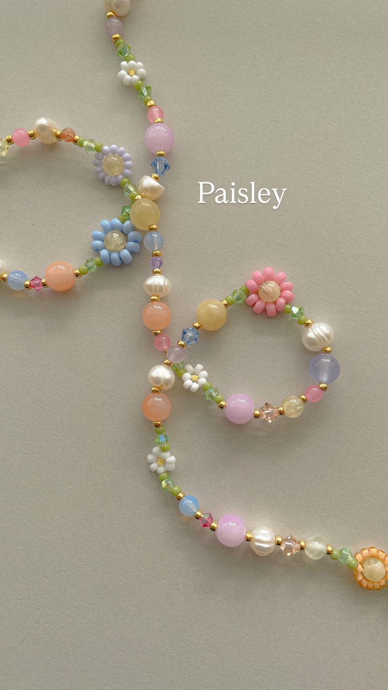 May include: A beaded necklace labeled Paisley featuring a variety of pastel-colored beads, including white pearls, translucent pink, blue, yellow, and purple spheres, interspersed with small flower-shaped beads in white, blue, and pink.