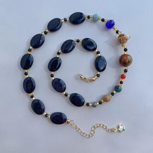May include: A necklace featuring oval dark blue goldstone beads interspersed with small gold-tone spacers and a series of multi-colored spherical beads representing planets. The piece includes a gold-tone chain extender with a small star charm.