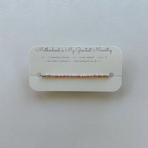 May include: A beaded bracelet with pink, gold, and gray beads is displayed on a white card. The card reads "Motherhood is My Greatest Ministry" in script, with a Morse code translation below.