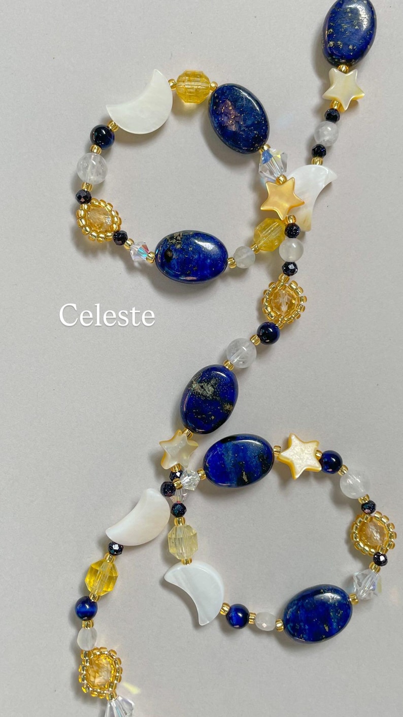 May include: A beaded necklace featuring oval lapis lazuli stones, white mother-of-pearl crescent moons, and star-shaped beads. The design includes yellow and clear faceted glass beads, small dark blue beads, and the word Celeste printed in white.